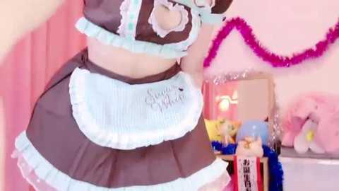 Media: Video of a person in a maid cosplay costume with frilly details, wearing a black and light blue apron. Background includes pink streamers and plush toys.