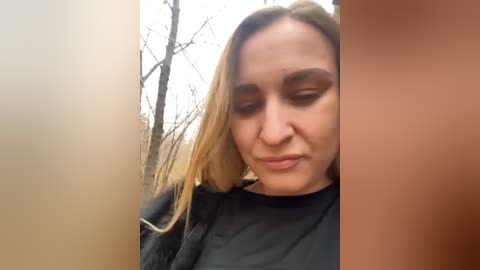 Media: A video of a young woman with shoulder-length blonde hair, wearing a black jacket, with a neutral expression, standing in a wooded area with bare trees in the background.