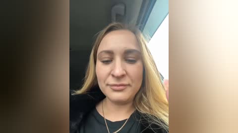 Media: A video of a young woman with light skin, long blonde hair, and light makeup, wearing a black jacket, sitting in a car with sunlight streaming in.