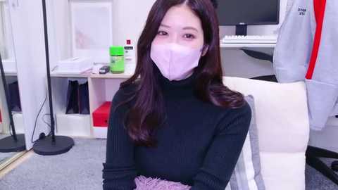 Media: Video of a young Asian woman with long dark hair, wearing a black turtleneck sweater, purple mask, and a fluffy pink blanket, seated in a modern office with white furniture, green chair, and computer setup.