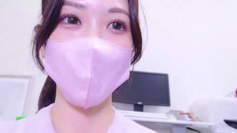 Media: Video of an Asian woman with light skin, dark hair, and large brown eyes wearing a pink surgical mask in a sterile medical office.