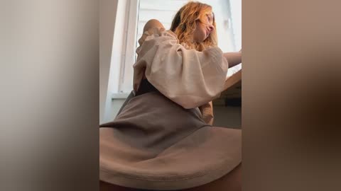 Media: Video of a woman with wavy, blonde hair, wearing a beige top and gray dress, leaning against a window, indoors.