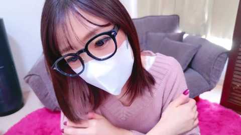 Media: Video of an Asian woman with shoulder-length brown hair, wearing black-rimmed glasses, white face mask, and a pink sweater. She is indoors, sitting on a pink rug, with a grey couch and curtains in the background.