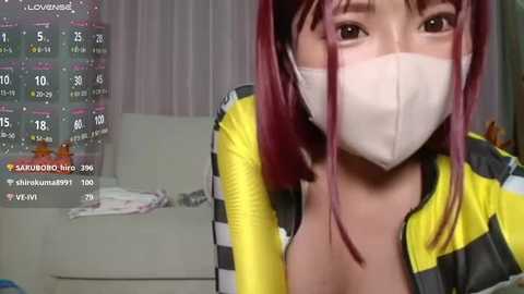 Media: A video of a young woman with fair skin and long, straight, red hair, wearing a yellow and gray striped hoodie, a white mask, and a gray sweatshirt. She's indoors, possibly in a bedroom with a bed and calendar in the background.