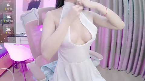 Media: Video of an East Asian woman with fair skin, wearing a white halterneck dress with a plunging neckline, revealing moderate cleavage, seated in a modern gaming chair with a computer setup in the background.