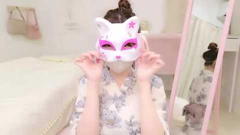 Media: Video of a young woman with pale skin, light brown hair in a bun, wearing a white floral kimono and a pink cat mask with white fur details. She stands in a pastel-colored room with a white bed and mirror.