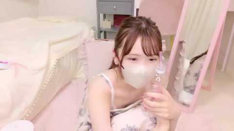 Media: Video of a young Asian woman in a pink floral dress with a face mask, kneeling and drinking from a bottle, in a pastel-colored, softly lit room with a white crib and pink furniture.