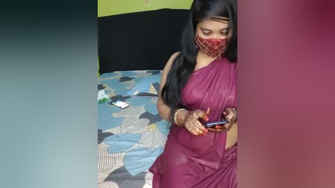 Media: Video of a woman in a red saree, mask, and bangles, hunched over a phone, in a simple bedroom with a patterned bedspread and green and black walls.