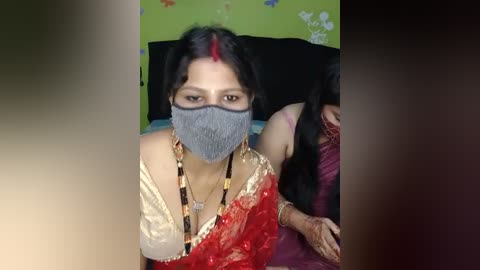 Media: Video of a South Asian woman with dark hair, wearing a red sari, gray mask, and beaded necklace, seated in a green and black room.