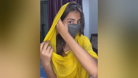 Media: Video of a young woman with medium brown skin, long black hair, wearing a bright yellow sari and a black face mask, adjusting her sari's pallu, in a dimly lit room with purple curtains.