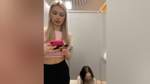 Media: Video of a blonde woman in a pink crop top and black leggings taking a selfie in a bathroom with a woman in the background, holding a tissue.