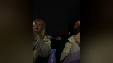 Media: Video of two people in a dimly lit car at night, with a blonde woman in a white coat and a man in a black hoodie and mask. A plastic water bottle is visible in the foreground.