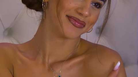 Media: Video of a smiling, fair-skinned woman with dark hair styled in an updo, wearing a strapless top, hoop earrings, and a delicate necklace.