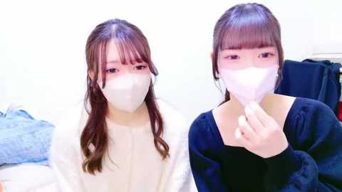 Media: Video of two young East Asian women with long, straight brown hair and bangs, wearing white surgical masks and white gowns, seated indoors with a blue blanket and dark clothing in the background.