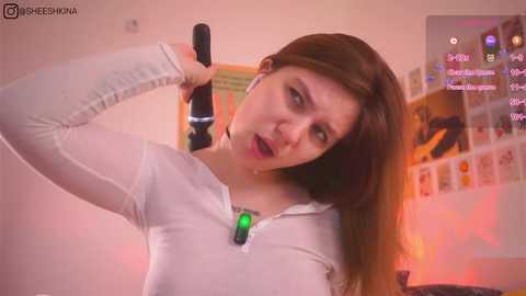 Media: Video of a young woman with straight brown hair, wearing a white long-sleeved shirt, holding a black hairdryer, in a cozy room with posters and a TV screen displaying a video game interface.