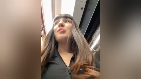 Media: Video of a young woman with long brown hair, straight bangs, and a black jacket, looking up in an indoor setting with fluorescent lighting.