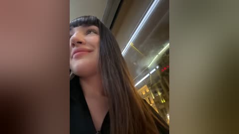 Media: Video of a smiling woman with long, straight brown hair and bangs, wearing a black top, taken indoors near a window with blurred city lights in the background.