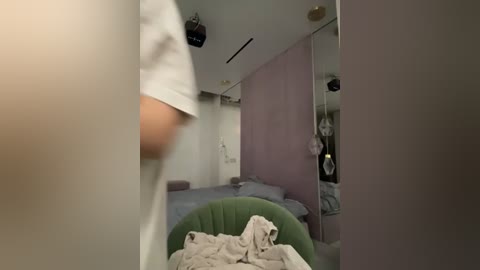 Media: Video of a modern, minimalist bedroom with light pink walls, a green beanbag chair, a messy bed, and a person in a white shirt walking away.