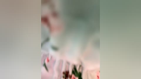 Media: A blurred video featuring a person with light skin, possibly a woman, wearing a floral dress. The background includes greenery and soft lighting, creating a serene, indistinct atmosphere.