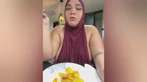 Media: Video of a young woman with medium skin tone and a maroon hijab, eating a plate of orange slices in a modern, dimly-lit room with hanging lights.