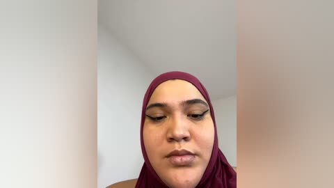 Media: Video of a young woman with light brown skin, wearing a burgundy hijab and subtle makeup, standing against a plain white wall. Her expression is neutral, eyes closed.