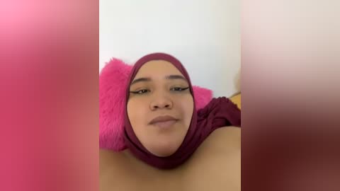 Media: Video of a woman with medium brown skin and a maroon hijab, lying on a bed with pink pillows, in a room with white walls. Her face is slightly blurred, focusing on her relaxed expression.