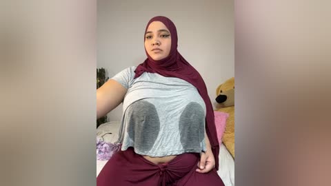 Media: Video of a plus-sized woman in a maroon hijab, gray crop top, and maroon pants, sitting on a bed with a teddy bear, in a cozy bedroom.