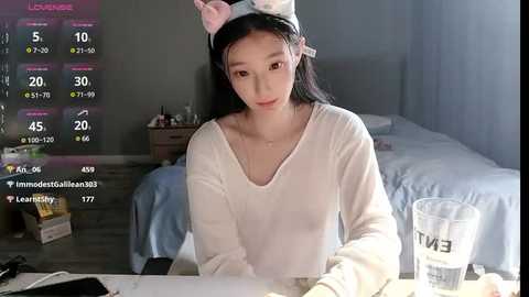 Media: A video of an Asian woman with long black hair and a pink cat headband, wearing a white long-sleeve shirt, sitting at a table in a dimly lit bedroom, with a glass of water and a phone in view.