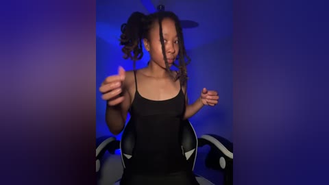 Media: Video of a young Black woman with curly hair in pigtails, wearing a black tank top, sitting on a gaming chair. The background is a gradient of blue and purple lights.