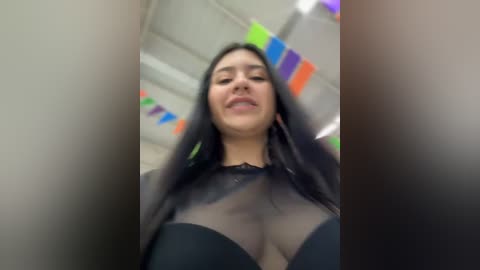 Media: A low-angle video captures a young woman with long black hair, fair skin, and a curvy physique, wearing a sheer black top revealing her large breasts. The background is blurred, featuring colorful streamers and an indoor setting.