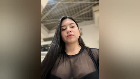 Media: Video of a young Latina woman with long black hair, wearing a sheer black top, standing indoors with a modern architectural background.