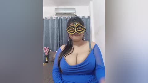 Media: A video of a plus-sized woman with medium skin tone, wearing a blue dress and a gold Venetian mask, standing in a room with gray curtains and a pink floral arrangement.