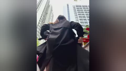 Media: A video captures a person walking on a city street, wearing a long black coat and backpack. Tall buildings and a green tree frame the background. The image is slightly blurred, emphasizing movement.