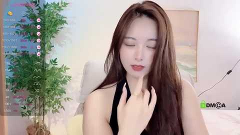 Media: A video of an Asian woman with long brown hair, wearing a black halter top, seated indoors with a lush green plant and abstract artwork in the background.