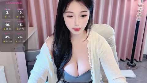 Media: A video of an East Asian woman with long black hair, wearing a revealing white cardigan over a gray bra, sitting at a desk in a pink-curtained room.