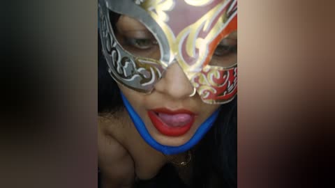 Media: Video of a close-up of a woman wearing a flamboyant, ornate Venetian masquerade mask with intricate silver designs, a bold blue chin strap, and striking red lipstick. Her dark hair peeks from beneath the mask.