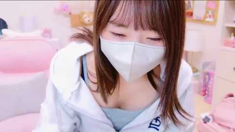 Media: Video of an Asian woman with straight brown hair, wearing a white face mask, light blue top, and white jacket, in a pastel-toned room with pink and white decor.