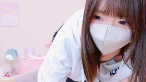 Media: Video of a young Asian woman with long brown hair and fair skin, wearing a white mask, white top, and gray pants, leaning forward in a bright, softly lit room with pink and white decor.