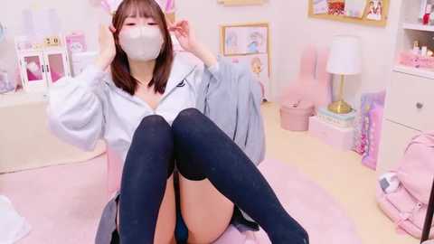 Media: Video of an Asian woman with straight brown hair, wearing a white mask, gray hoodie, blue skirt, and black thigh-high socks, seated in a pastel-themed, cluttered room with pink furniture and toys.