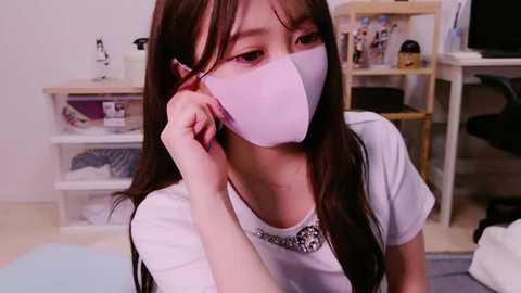 Media: Video of an Asian woman with long brown hair, wearing a white mask, white shirt, and silver necklace, seated indoors with shelves and office equipment in the background.