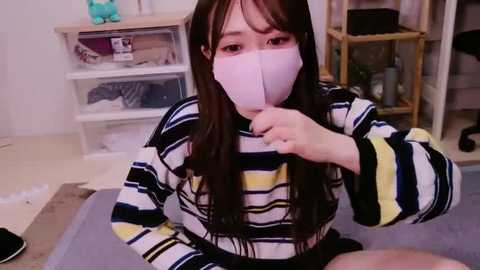 Media: Video of an Asian woman with long brown hair, wearing a pink face mask and a striped sweater, seated indoors with a wooden shelf in the background.