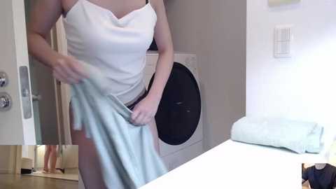 Media: Video of a woman in a white tank top and shorts, pulling a light blue towel from a dryer in a modern, bright laundry room with white walls and a door.