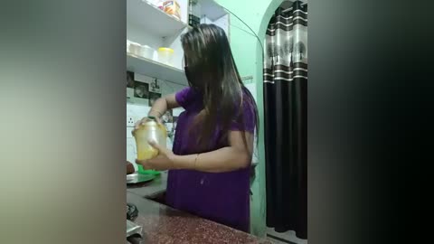 Media: Video of a woman in a purple shirt pouring liquid from a jar into a container in a kitchen with green walls and a striped curtain.