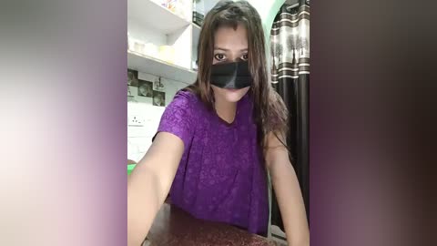 Media: Video of a young woman with long brown hair, wearing a black face mask, a purple lace top, and a red patterned skirt, sitting in a cluttered bathroom with white tiles and a striped curtain.