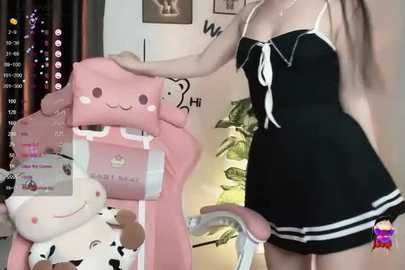 Media: Video of a young woman with light skin, wearing a black and white striped maid outfit, sitting on a pink, plush chair. Background shows a cozy room with framed pictures and a green plant.