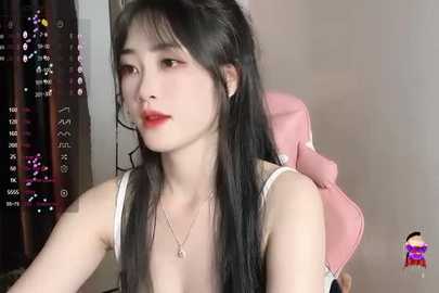 Media: Video of an East Asian woman with long, straight black hair, fair skin, red lipstick, and a white tank top, sitting in a pink gaming chair. Background shows a dark curtain with a digital clock.