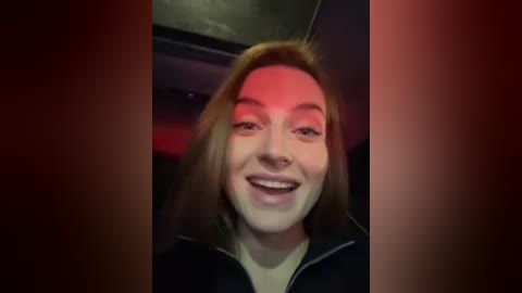 Media: Video of a young woman with fair skin, light brown hair, and a bright smile. She's wearing a black jacket. The background is dimly lit with red and blue lighting.