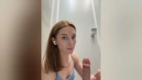 Media: Video of a fair-skinned woman with straight brown hair, wearing a light blue top, standing in a white hallway. She has a neutral expression and holds a large, erect penis in her right hand.