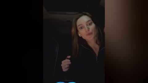 Media: Video of a Caucasian woman with long brown hair, wearing a black hoodie, sitting in a dimly lit car, with her left hand near her mouth.