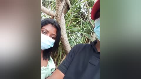 Media: A video of a woman with a light complexion, wearing a blue surgical mask, green top, and short black hair, standing behind a man in a black polo shirt and red hat, surrounded by lush green palm leaves.